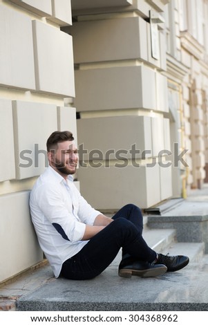 Close-up portrait of man freelancer sitting and waiting near building in the city centre. Bearded man in white shirt and black jeans smiling outdoors.