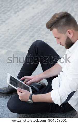 Technology, lifestyle and business concept. Handsome man freelancer working with tablet pc oudoors. Man in black jeans and white shirt.