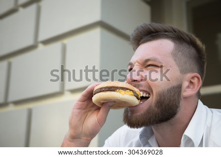 Man eating hot hamburger after stressful work. Close-up of hipster bearded man eating delicious hamburger outdoors.