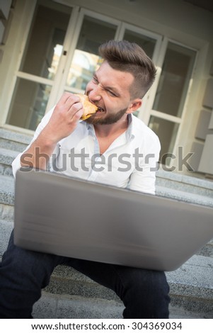 Man freelancer eating hamburger with laptop on his knees. Man both working and eating. He looks so hungry during his work.