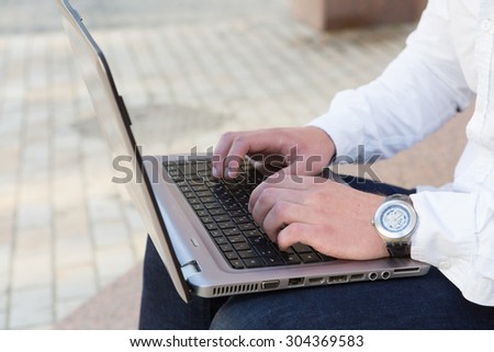 Cropped shot of businessman\'s hands typing on laptop that is on his knees. Man in business suit sitting outside with expensive watch on his wrist.