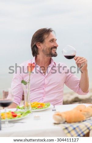 Handsome man drinking red wine in the restaurant by the sea. Bearded man in pink shirt relaxing and enjoying daytime.