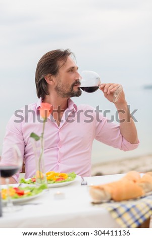 Portrait of handsome man drinking red wine. Bearded man in pink shirt sitting at the table in the cafe near by sea.