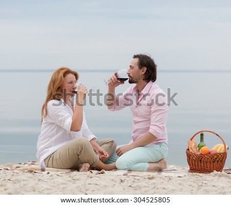 holidays, vacation and happy people concept. People having picnic near by sea in the vening. People drinking red wine, basket is near them.