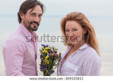 Smiling couple spending their time near the ocean. Man in pink shirt and woman in white shirt posing with bouquet of flowers for the camera.