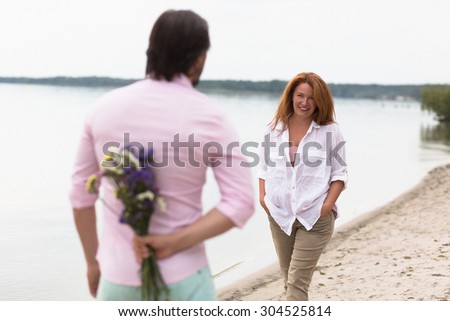 Romantic couple walking on sandy beach. Man hiding bouquet of flowers for his wife. Red-haired woman reaching to him with hands in pockets.