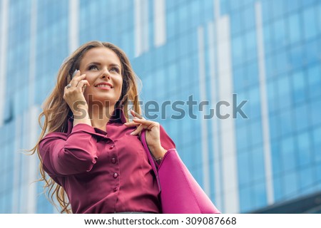 Businesswoman speaking over mobile phone and walking near office building. Pretty lady holding vivaceous bag at her hand in the city.