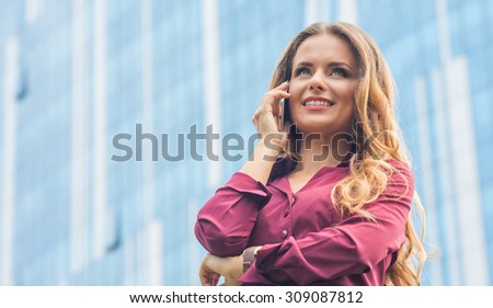 Businesswoman working with her partners in the city over mobile phone. Woman in vinaceous blouse standing near office building.