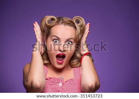 Surprised young girl with her hands raised in photo studio. Astonished pin-up girl with blond hair screaming or shouting.