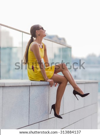 Fashion girl in little yellow dress sitting on brick wall with crossed legs. Pretty lady in sunglasses and high heels looking away.