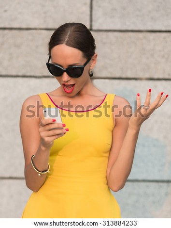 Fashion girl in yellow dress looking at mobile phone's screen with amazement. Surprised lady in sunglasses standing near brick wall.
