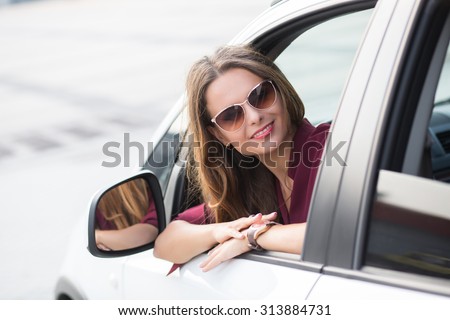 Portrait of young businesswoman sitting in car and looking from it. Lady in sunglasses smiling and looking at the camera.