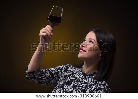 Smiling asian sommelier woman raised a glass of red wine to study it colour. Beautiful middle-aged woman in traditional dress posing over dark background.