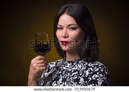 Close-up portrait of asian woman holding a glass of red wine near her face. Middle-aged woman with red lips posing for photographer.
