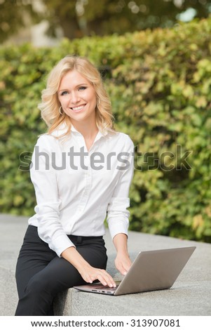 Freelance lady working with laptop computer and smiling for the camera oudoors. Blonde in white shirt and black trousers sitting on brick wall.