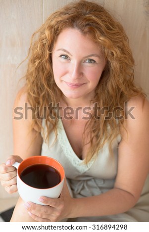 Close-up portrait of middle-aged woman drinking hot black tea. Happy lady with red hair smiling for the carema at her home.