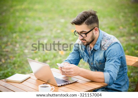 Serious freelancer hipster man using his smart phone. Handsome man working with his computer in the park.