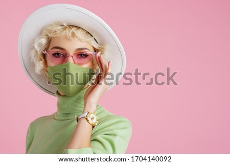 Woman wearing trendy fashion outfit during quarantine of coronavirus outbreak. Model dressed protective stylish handmade face mask, pink sunglasses, white hat, wrist watch, green mint color turtleneck