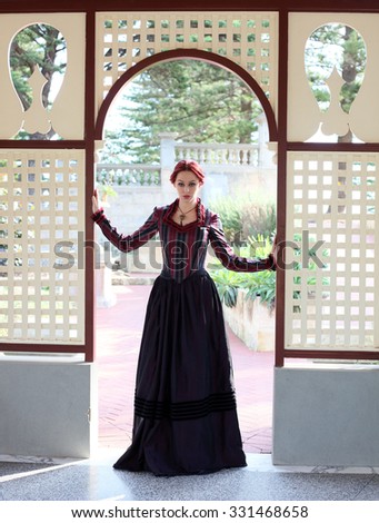 Full Length Portrait of a beautiful red haired girl wearing  Gothic inspired Victorian era clothes. standing in a window frame, looking at camera.