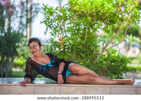 Attractive elegant young woman in bikini laying down on stone bench,beautiful green bush bokeh behind her