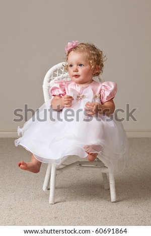 One year old baby girl wearing white and pink dress sitting in a white wicker chair.