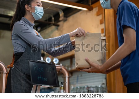 Young Asian restaurant owner or waitress wearing face mask giving takeaway food bag to delivery man. Male courier taking online order for delivery at restaurant. Takeaway food during covid-19 pandemic