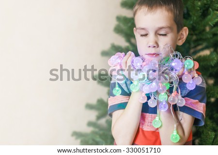 make a wish under Christmas. beautiful boy covering his eyes with long eyelashes make a wish. He holds in his hands and chest presses to Christmas lights