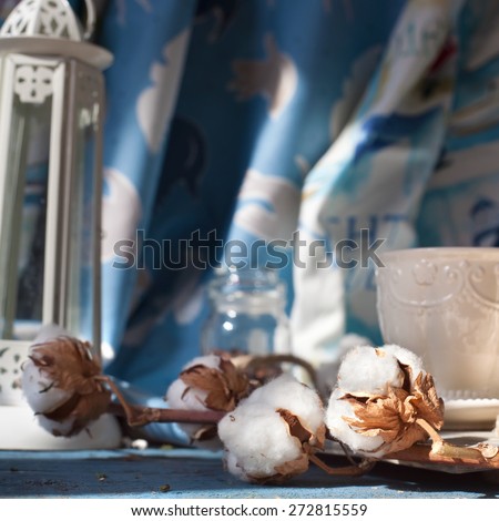 Branch of cotton flowers set with home decorations: cotton fabrics, cup of coffee and a lighthouse candle holder, shot in natural daylight. Marine style home decorations and sea holiday concept.