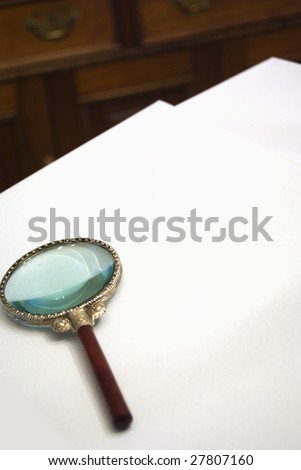 Magnifying glass on blank paper with desk in background.  Customization space for text or document insert.