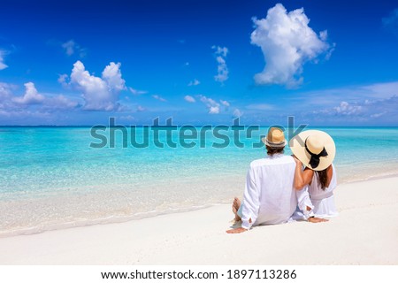 A beautiful tourist couple with hats sits on a tropical beach and enjoys the view to the turquoise ocean during their summer holiday