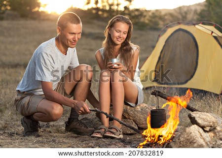 Young couple sitting near tent and looking at the fire while water is boiling