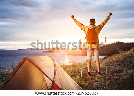 Backpacker with arms raised looking at beautiful sunrise at big lake near tent