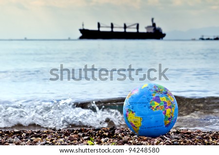 THE AQABA GULF, RED SEA – February 03, 2012: A small color globe of earth on marine beach with a cargo ship on horizon and crushing wave nearly are symbols of   ecological fragility of our planet