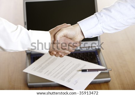 Business handshake after the signing of a contract with laptop and contract in background.