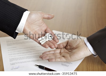one hand of businessman gives  house keys to the hand of another businessman. Signed contract and pen visible in background.