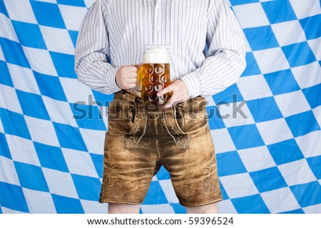 Bavarian man with Oktoberfest beer stein (Mass) and leather pants (Lederhose). In background is Bavarian flag visible.