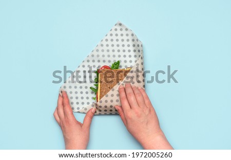 Woman's hands wrapping a sandwich in a beeswax cloth on a blue table, above view. Healthy sandwich with wholemeal bread and vegan ingredients, wrapped in reusable beeswax paper.