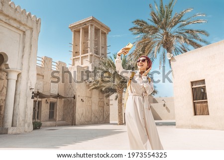 Happy woman wearing a turban hat takes a selfie photo on her smartphone against the background of a Bur Dubai old town near Creek district