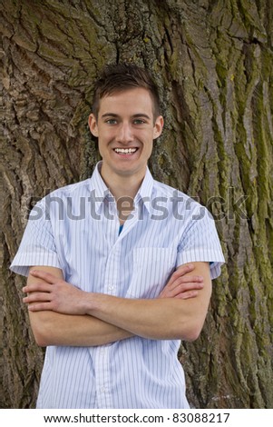 smiling fashionable young man standing next to a tree and posing with his arms crossed