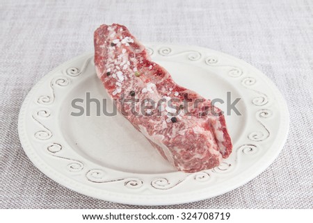 A piece of fresh marbled beef with sea salt and black pepper on the porcelain plate.