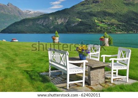 it is a garden place of a private cottage in the border of a fijord: the grass with a table and chairs decorated with flowers, beside fjord are mountains covered with green woods, and snow in top