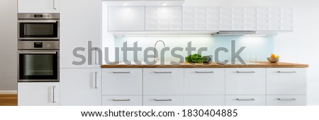 Panorama of modern kitchen with white furniture, wooden countertop and build in fridge, oven and microwave