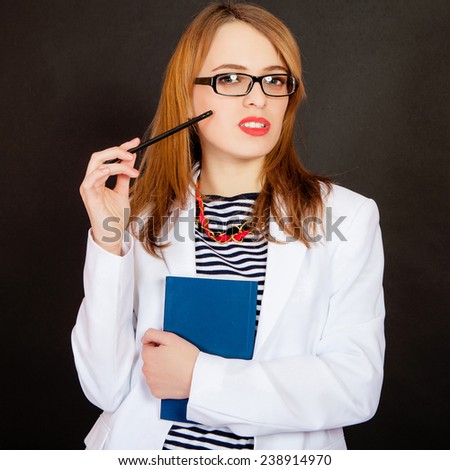 Fashionable doctor. Confident young female doctor in white uniform and glasses holding book and pencil while standing against black background