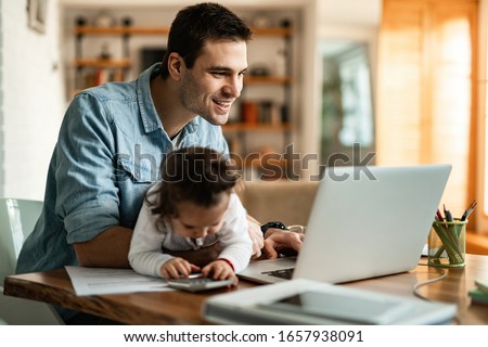 Photo of Young happy father reading e-mail on a computer while being with his baby daughter at home. 