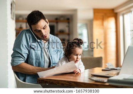 Photo of Young working father babysitting his small daughter and making a phone call at home. 