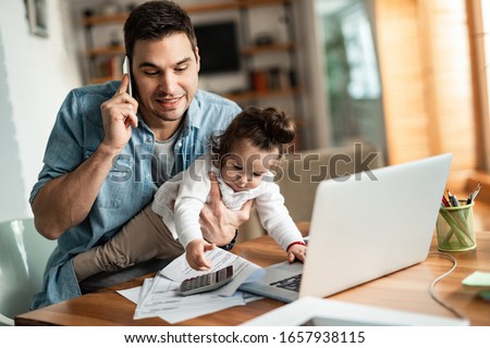 Photo of Young working father talking on the phone while babysitting his playful daughter at home.