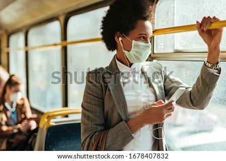 Black businesswoman with protective face mask using smart phone and looking through the window while commuting by bus.  
