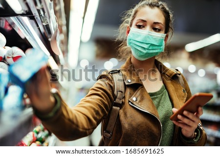 Young woman with face mask using mobile phone and buying groceries in the supermarket during virus pandemic. 