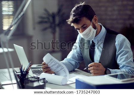 Male entrepreneur analyzing business reports while wearing face mask and working in the office during virus epidemic. 