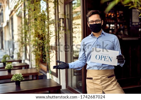 Happy waiter with protective face mask holding open sign while standing at cafe doorway. 